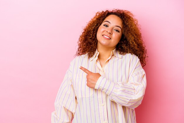 Joven mujer latina con curvas aislada en la pared rosa sonriendo y apuntando a un lado, mostrando algo en el espacio en blanco.