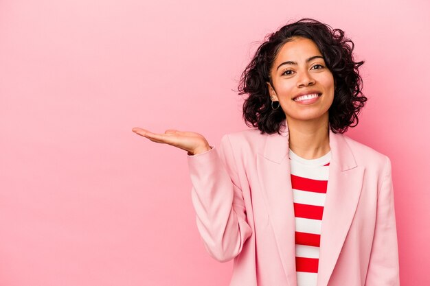 Joven mujer latina de moda aislada sobre fondo rosa mostrando un espacio de copia en una palma y sosteniendo otra mano en la cintura.