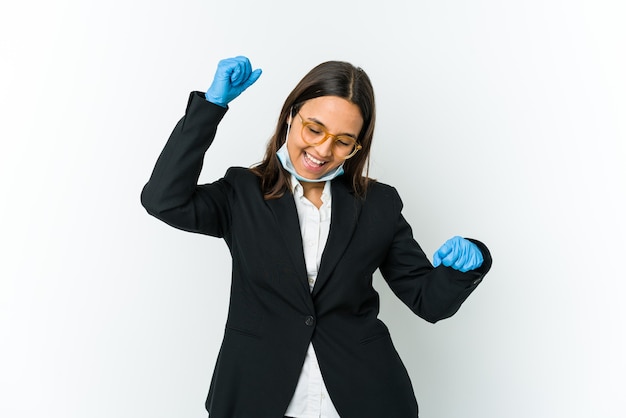 Foto joven mujer latina de negocios con una máscara para protegerse del covid aislado en la pared blanca celebrando un día especial, salta y levanta los brazos con energía.