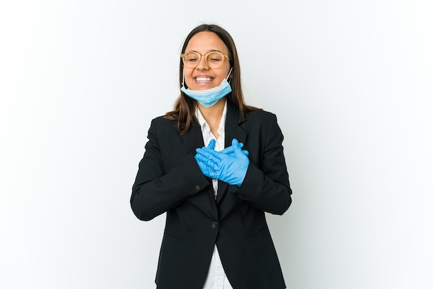 Joven mujer latina de negocios con una máscara para protegerse del covid aislado sobre fondo blanco riendo manteniendo las manos en el corazón, concepto de felicidad.