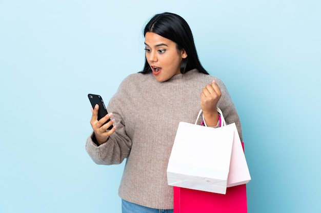 Joven mujer latina en la pared azul con bolsas de compras y escribir un mensaje con su teléfono celular a un amigo