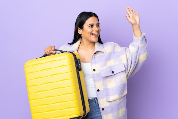 Joven mujer latina en pared púrpura en vacaciones con maleta de viaje y saludando