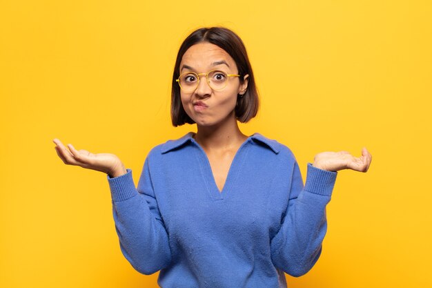 Joven mujer latina que se siente perpleja y confundida, dudando, ponderando o eligiendo diferentes opciones con expresión divertida