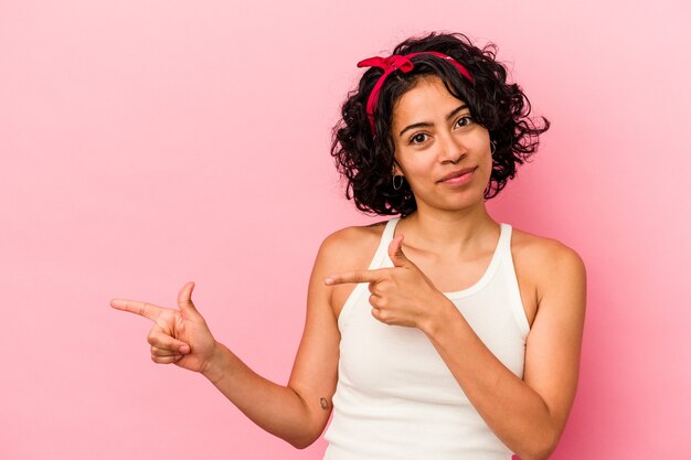 Joven mujer latina rizada aislada sobre fondo rosa sorprendido apuntando con los dedos índices a un espacio de copia.