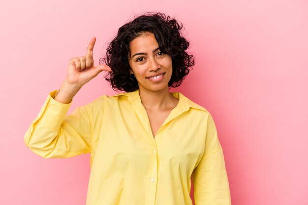 Joven mujer latina rizada aislada sobre fondo rosa sosteniendo algo pequeño con los dedos índices, sonriendo y confiado.