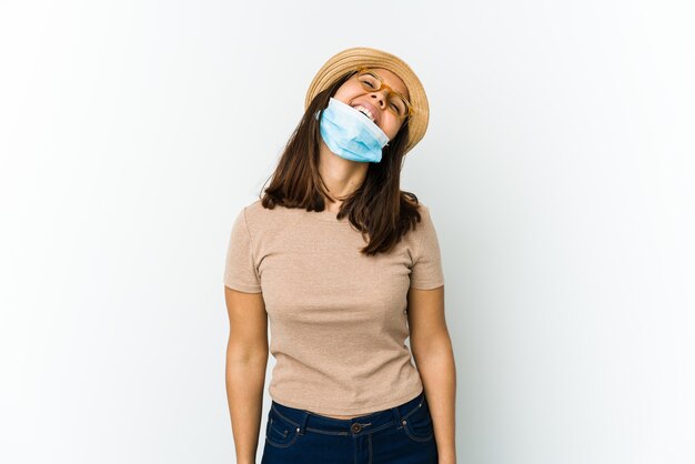 Joven mujer latina con sombrero y máscara para protegerse del covid aislado en la pared blanca riendo relajado y feliz, cuello estirado mostrando los dientes.