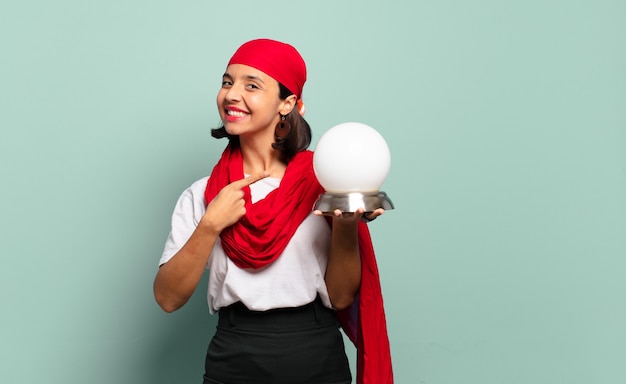 Joven mujer latina sonriendo alegremente, sintiéndose feliz y apuntando hacia un lado y hacia arriba, mostrando el objeto en el espacio de la copia