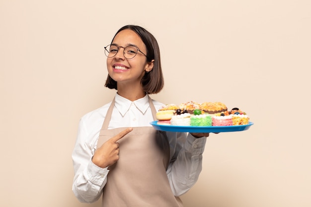 Joven mujer latina sonriendo alegremente, sintiéndose feliz y apuntando hacia un lado y hacia arriba, mostrando el objeto en el espacio de la copia