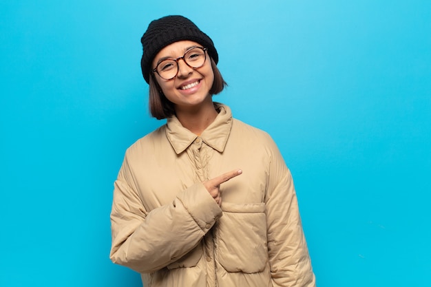 Joven mujer latina sonriendo alegremente, sintiéndose feliz y apuntando hacia un lado y hacia arriba, mostrando el objeto en el espacio de la copia