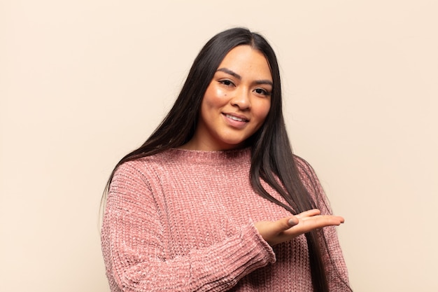 Joven mujer latina sonriendo alegremente, sintiéndose feliz y mostrando un concepto en el espacio de la copia con la palma de la mano