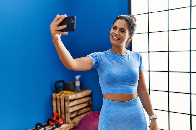 Foto joven mujer latina sonriendo confiada se hace selfie con el teléfono inteligente en el centro deportivo