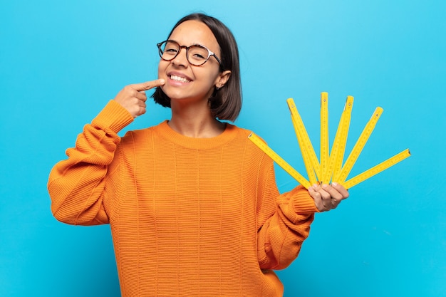 Joven mujer latina sonriendo con confianza apuntando a su propia sonrisa amplia, actitud positiva, relajada y satisfecha