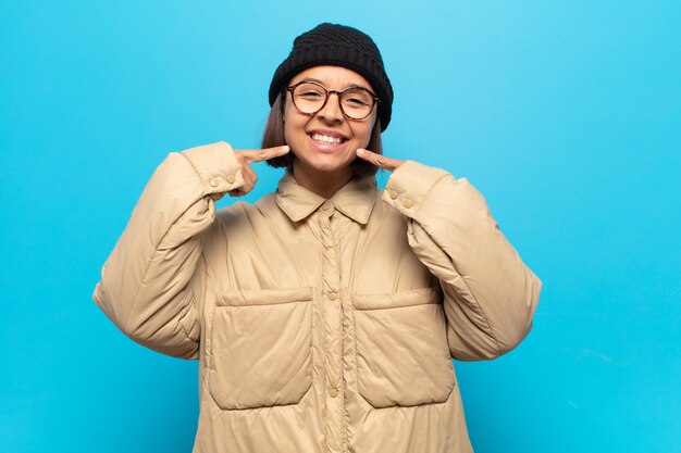 Joven mujer latina sonriendo con confianza apuntando a su propia sonrisa amplia, actitud positiva, relajada y satisfecha