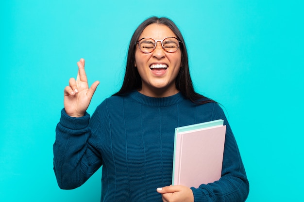 Joven mujer latina sonriendo y cruzando ansiosamente ambos dedos, sintiéndose preocupada y deseando o esperando buena suerte