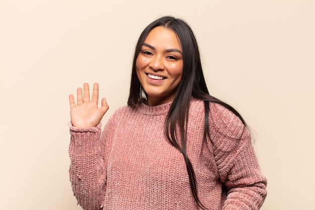 Joven mujer latina sonriendo feliz y alegremente, saludando con la mano, dándote la bienvenida y saludándote, o diciéndote adiós