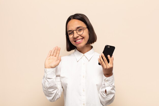 Joven mujer latina sonriendo feliz y alegremente, saludando con la mano, dándote la bienvenida y saludándote, o diciéndote adiós