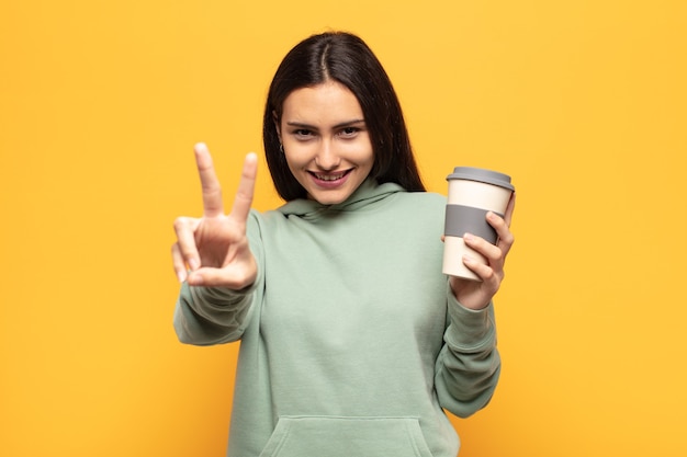 Joven mujer latina sonriendo y luciendo feliz, despreocupada y positiva, gesticulando victoria o paz con una mano