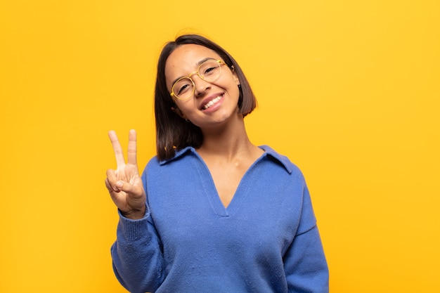 Joven mujer latina sonriendo y luciendo feliz, despreocupada y positiva, gesticulando victoria o paz con una mano