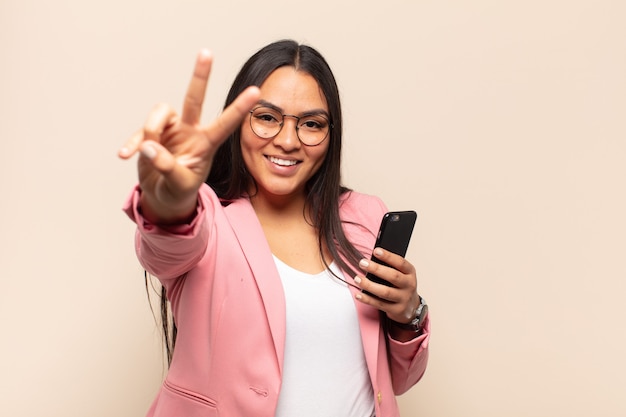 Joven mujer latina sonriendo y luciendo feliz, despreocupada y positiva, gesticulando victoria o paz con una mano