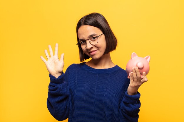 Joven mujer latina sonriendo y mirando amistosamente, mostrando el número cinco o quinto con la mano hacia adelante, contando hacia atrás