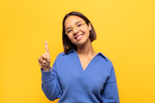 Joven mujer latina sonriendo y mirando amistosamente, mostrando el número uno o el primero con la mano hacia adelante, contando hacia atrás