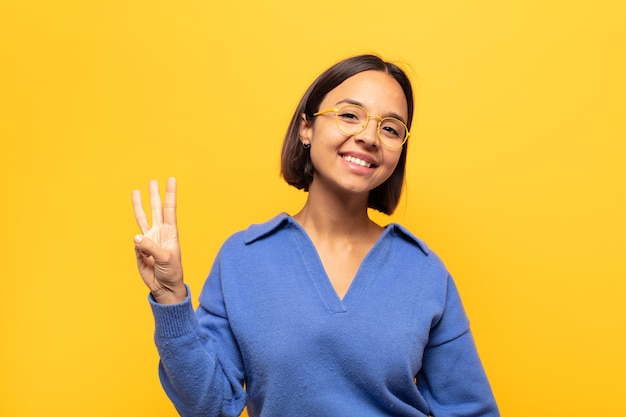 Joven mujer latina sonriendo y mirando amistosamente, mostrando el número tres o tercero con la mano hacia adelante, contando hacia atrás