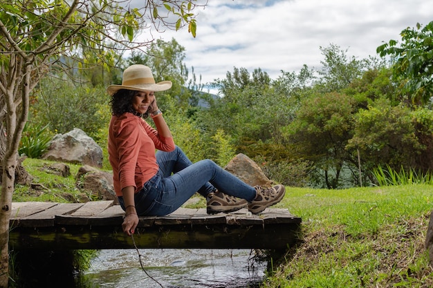 Joven mujer latina sonriente con sombrero sentado en un viejo puente de madera sobre un pequeño arroyo