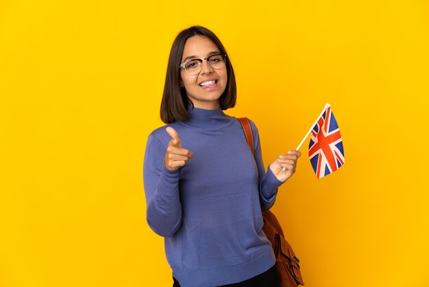 Joven mujer latina sosteniendo una bandera del Reino Unido aislada en la pared amarilla apuntando hacia el frente y sonriendo
