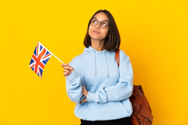 Joven mujer latina sosteniendo una bandera del Reino Unido aislada en la pared amarilla pensando en una idea mientras mira hacia arriba