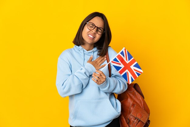 Joven mujer latina sosteniendo una bandera del Reino Unido aislado sobre fondo amarillo con un dolor en el corazón