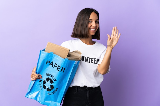 Joven mujer latina sosteniendo una bolsa de reciclaje llena de papel para reciclar aislado en morado saludando con la mano con expresión feliz