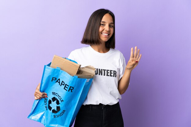 Joven mujer latina sosteniendo una bolsa de reciclaje llena de papel para reciclar aislado en púrpura feliz y contando cuatro con los dedos