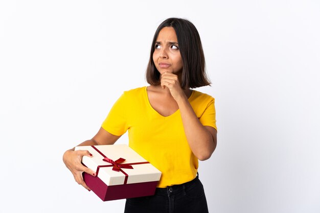 Joven mujer latina sosteniendo un regalo aislado teniendo dudas y pensando