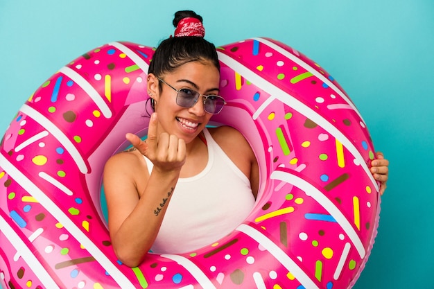 Joven mujer latina sosteniendo una rosquilla inflable aislada sobre fondo azul apuntando con el dedo hacia usted como si estuviera invitando a acercarse.