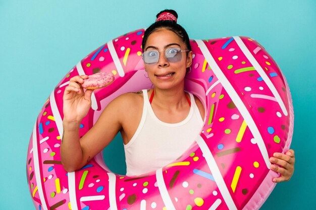 Joven mujer latina sosteniendo una rosquilla inflable comiendo una rosquilla aislado sobre fondo azul.