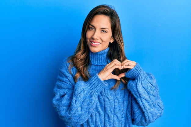 Joven mujer latina con suéter casual de invierno sonriendo enamorada haciendo forma de símbolo de corazón con las manos. concepto romántico.