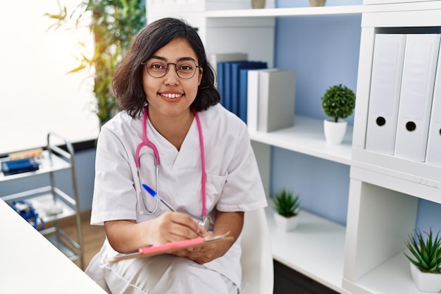 Joven mujer latina con uniforme médico escribiendo en el portapapeles en la clínica