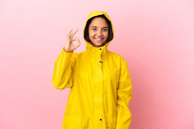 Joven mujer latina vistiendo un abrigo impermeable sobre antecedentes aislados que muestra el signo de ok con los dedos