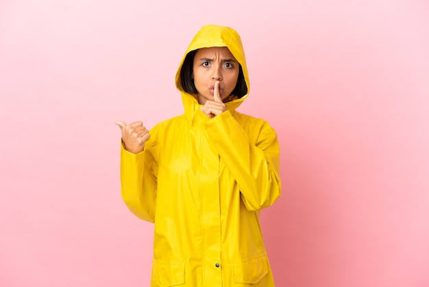 Joven mujer latina vistiendo un abrigo impermeable sobre fondo aislado apuntando hacia el lado y haciendo gesto de silencio