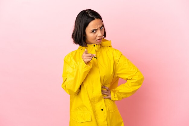 Joven mujer latina vistiendo un abrigo impermeable sobre fondo aislado frustrado y apuntando hacia el frente