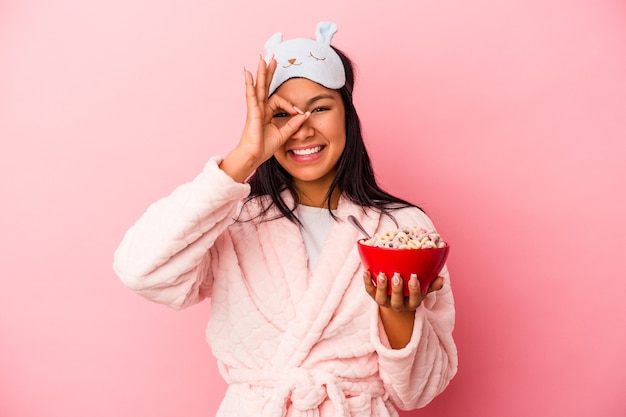 Joven mujer latina vistiendo pijama sosteniendo un tazón de cereales aislado sobre fondo rosa emocionado manteniendo el gesto ok en el ojo.