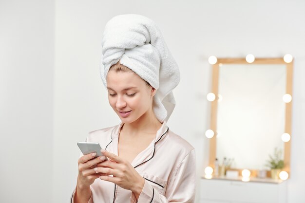 Foto joven mujer limpia con una toalla blanca suave en la cabeza desplazándose a través de mensajes en el teléfono inteligente después de la ducha matutina