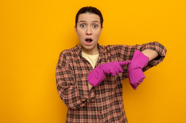 Joven mujer de la limpieza en camisa a cuadros con guantes de goma confundida apuntando a su brazo recordando el tiempo de pie sobre la pared naranja