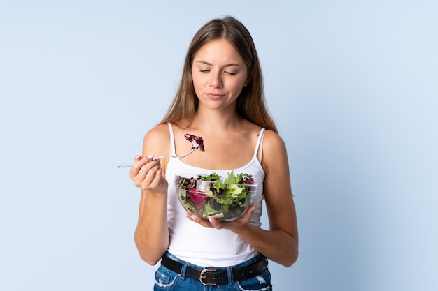 Joven mujer lituana aislada en azul sosteniendo un plato de ensalada con expresión triste