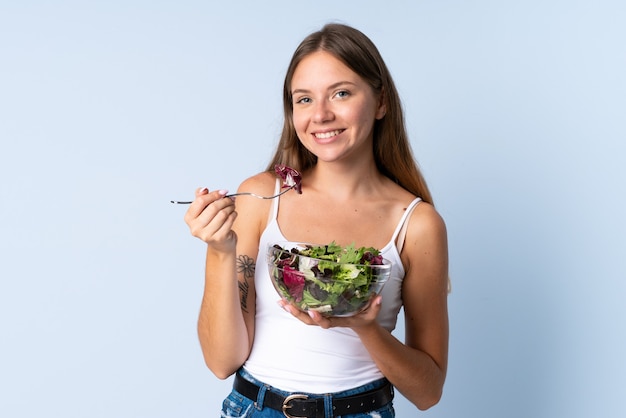 Joven mujer lituana aislada sobre fondo azul sosteniendo un plato de ensalada con expresión feliz