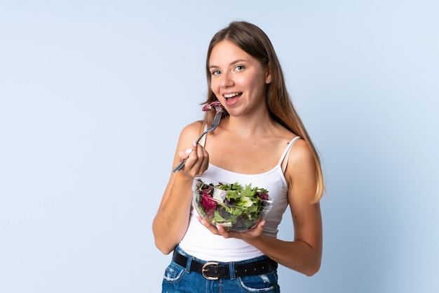 Joven mujer lituana aislada sobre fondo azul sosteniendo un plato de ensalada con expresión feliz