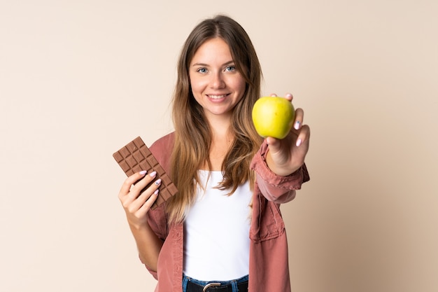 Joven mujer lituana aislada sobre fondo beige tomando una tableta de chocolate en una mano y una manzana en la otra