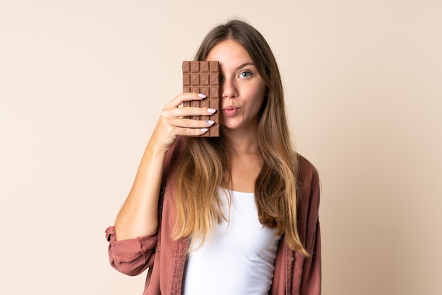 Foto joven mujer lituana aislada sobre fondo beige tomando una tableta de chocolate y sorprendida