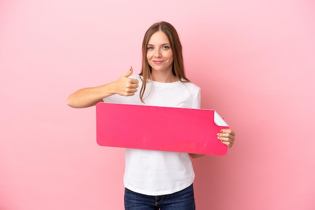 Joven mujer lituana aislada sobre fondo rosa sosteniendo un cartel vacío con el pulgar hacia arriba