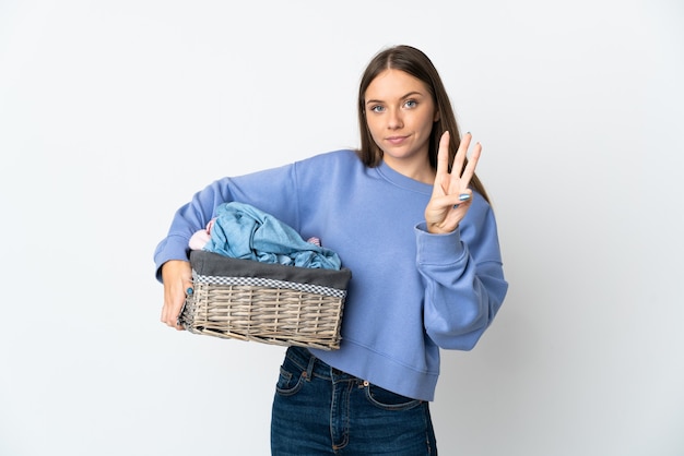 Joven mujer lituana sosteniendo una canasta de ropa aislada en la pared blanca feliz y contando tres con los dedos
